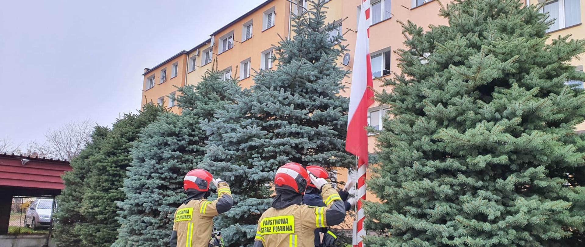 Zdjęcie przedstawia 3 strażaków stojących do nas tyłem
z podniesionymi palcami przyłożonymi do hełmu w geście
salutowania, wszyscy są ubrani w ubrania specjalne
dwóch z nich po bokach ma ubrania specjalne
w kołorze piaskowym
jeden który stoi obok masztu ma ubranie specjalne koloru czarnego i wciąga
flabę Polski na masz
tuż za nimi widać rosnące choinki a tuż za choinkami \
w oddali jest blok
po lewej stronie można dostrzec wiatę garażową a tuż za nią s
samochód osobowy koloru srebrnego.