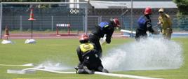 Dzień. Na murawie boiska sportowego trzech druhów ubranych ubranych w czarne ubranie koszarowe i czerwone hełmy rozwija linie gaśnicze. Jeden ze strażaków klęczy na wężu strażackim z którego tryska duży strumień wody. Obok druhów stoi funkcjonariusz PSP ubrany w mundur służbowy w kolorze piaskowym, który pełni rolę sędziego. W tle widać bramkę boiska sportowego oraz bieżnie.