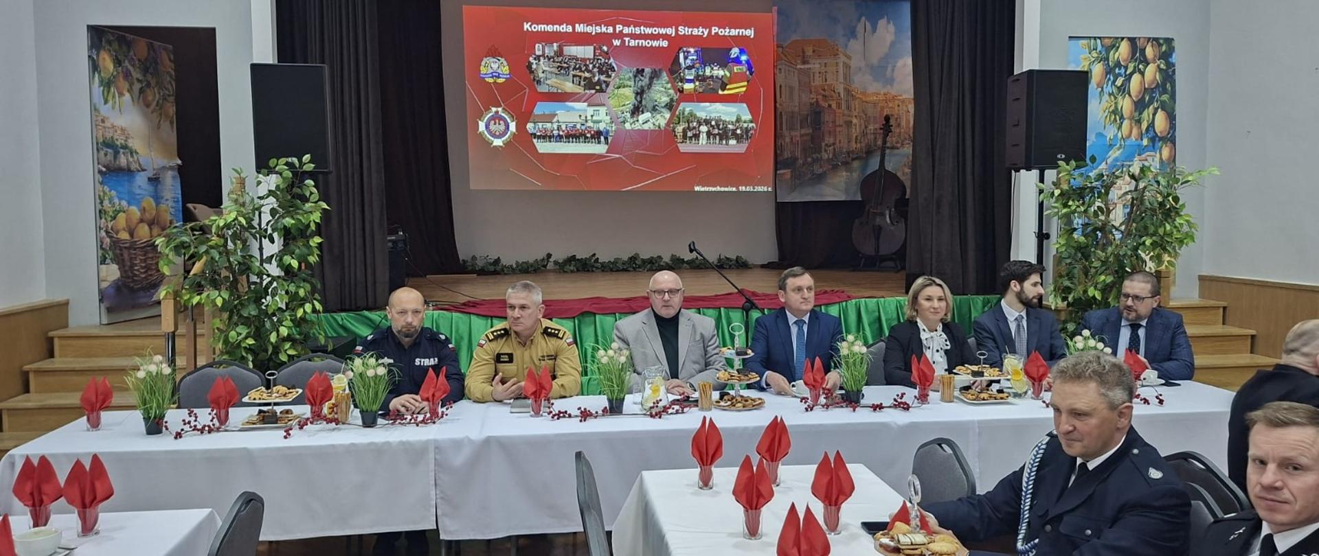 Przy długim stole siedzą przedstawiciele straży pożarnej oraz zaproszeni goście w strojach służbowych i formalnych. W tle widoczna scena z ekranem prezentacyjnym wyświetlającym slajd dotyczący działalności Komendy Miejskiej Państwowej Straży Pożarnej w Tarnowie.