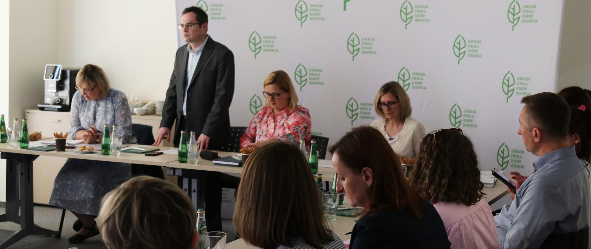 widok na uczestników spotkania, część z nich siedzi za stołem konferencyjnym, mężczyzna stoi i wita gości. Za nim widać białą ściankę reklamową z powielonym zielonym logotypem Generalnej Dyrekcji Ochrony Środowiska (liść)