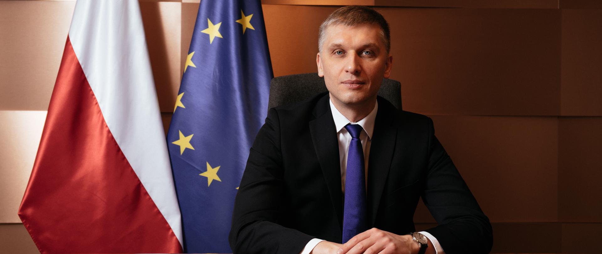 Na zdjęciu Minister Rozwoju i Technologii Piotr Nowak - siedzi za stołem, patrzy w obiektyw, w tle flagi Polski i Unii Europejskiej