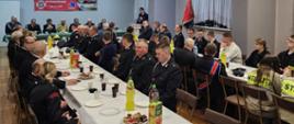 Sala wiejska w Rogożewie. Na pierwszym planie stoły nakryte białymi obrusami, na których znajdują się talerze z ciastem, kubki i napoje. Przy stołach zasiadają uczestnicy zebrania. W tle ustawiony jest stół prezydialny. Za stołem zasiadają goście oraz przedstawiciele zarządu OSP. Z prawej strony stołu prezydialnego, przy mównicy stoi przewodniczący zebrania, który przemawia do zebranych. 