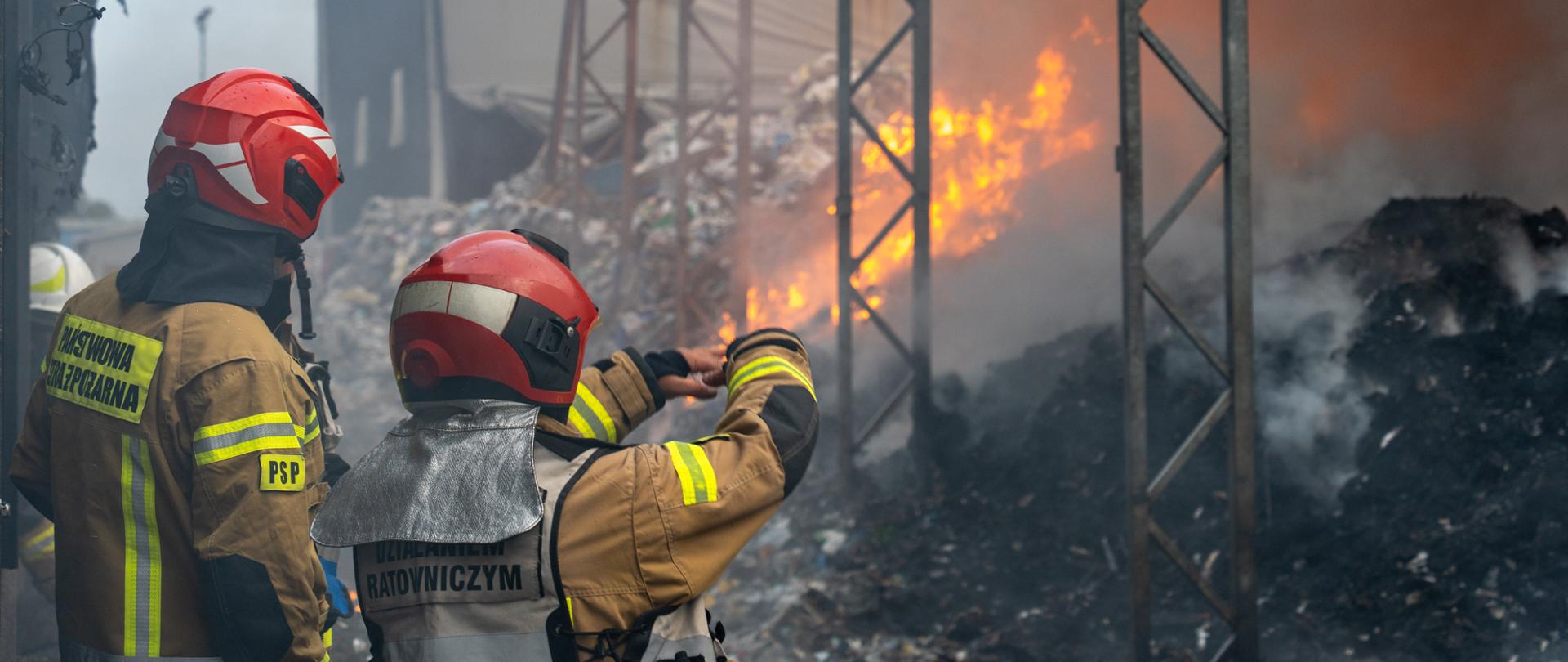 Na zdjęciu widać dwóch strażaków w ubraniach specjalnych i hełmach, jeden z nich ma biało-niebieską kamizelkę z napisem „Kierujący Działaniem Ratowniczym”, w tle objęta pożarem pryzma śmieci pod zadaszeniem