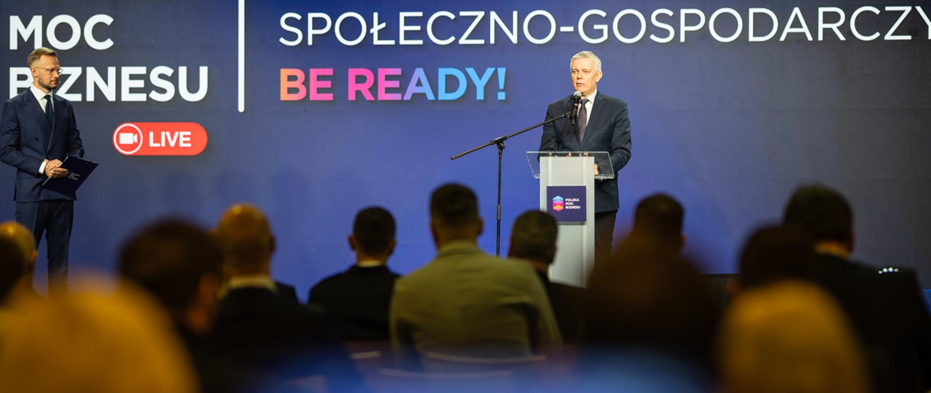 Mężczyzna za mównicą przemawia do zgromadzonych na sali.