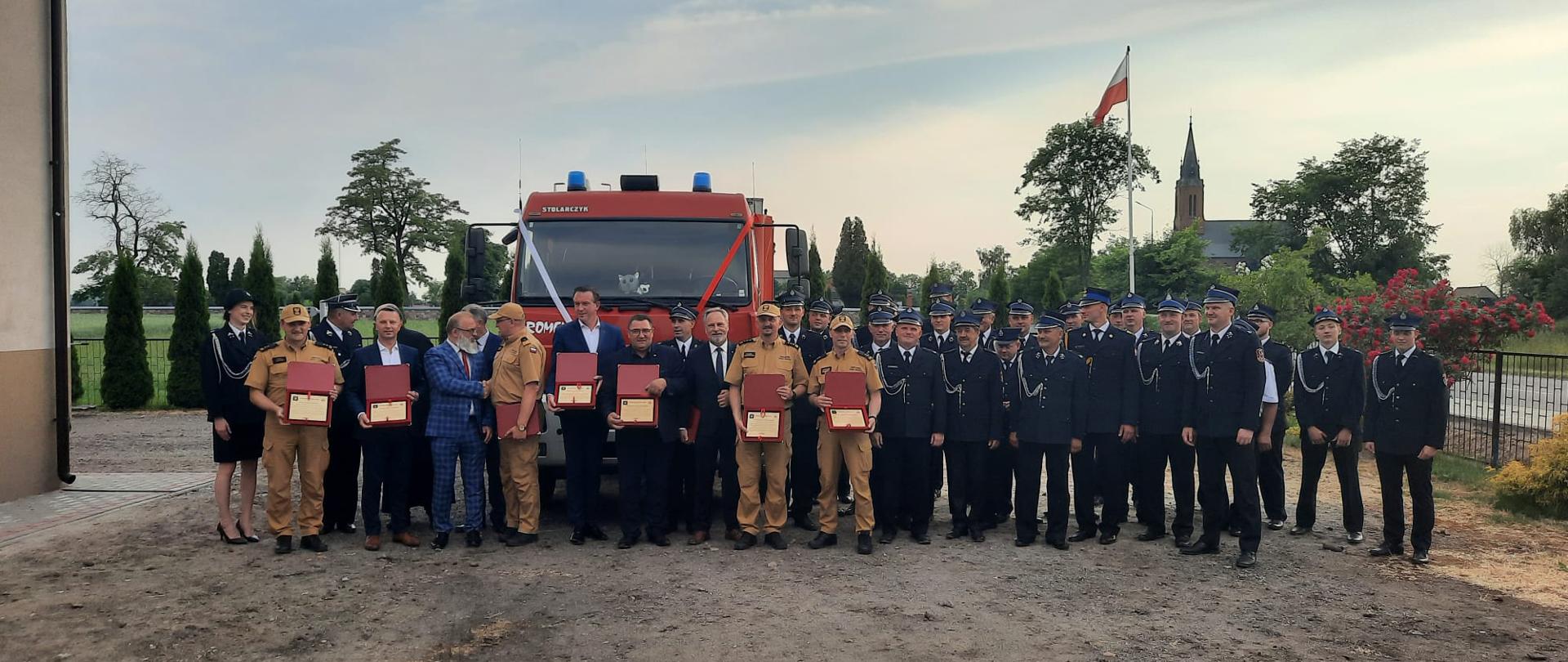 druhowie OSP, strażacy oraz goście uroczystości pozują do zdjęcia na tle samochodu ratowniczo-gaśniczego, w tel drzewa i wieża kościelna