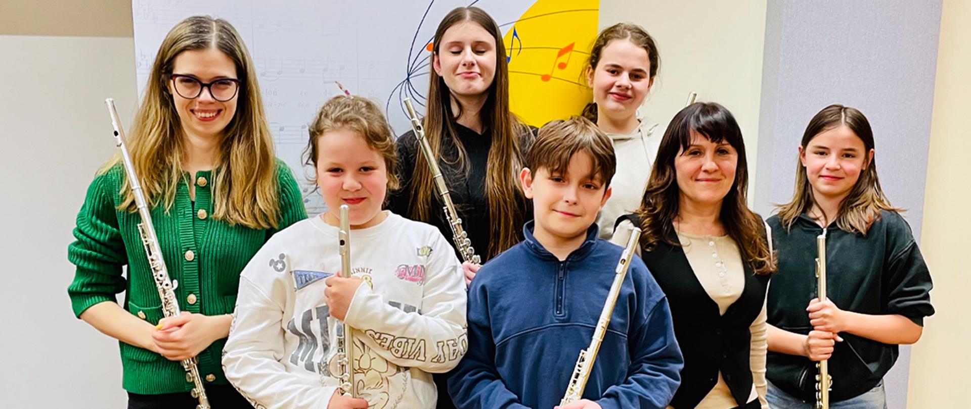 Na zdjęciu dyrektorka szkoły, nauczycielka prowadząca warsztaty i pięcioro uczniów stoją w auli szkoły na tle baneru, w rękach trzymają flety.