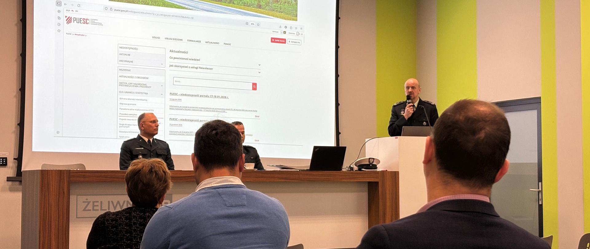Zdjęcie przedstawia spotkanie o charakterze urzędowym. W sali konferencyjnej widać prelegenta w mundurze (inspektor Inspekcji Transportu Drogowego), stojącego przy mównicy i przemawiającego do uczestników. Obok, za stołem, siedzi jeszcze dwóch inspektorów.
Na dużym ekranie za nimi wyświetlona jest prezentacja – strona internetowa PUESC (Platforma Usług Elektronicznych Skarbowo-Celnych). Na pierwszym planie widać uczestników siedzących tyłem do aparatu i słuchających prezentacji. Sala jest nowoczesna, z jasnymi ścianami i zielonymi akcentami, a całość sprawia wrażenie formalnego, oficjalnego wydarzenia szkoleniowego.