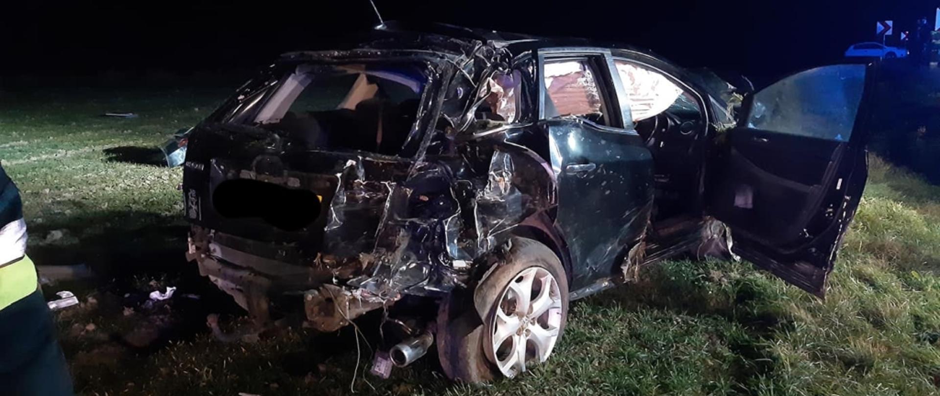 Na zdjęciu widoczny rozbity samochód osobowy w ciemnym kolorze, stojący na trawie, poza jezdnią.