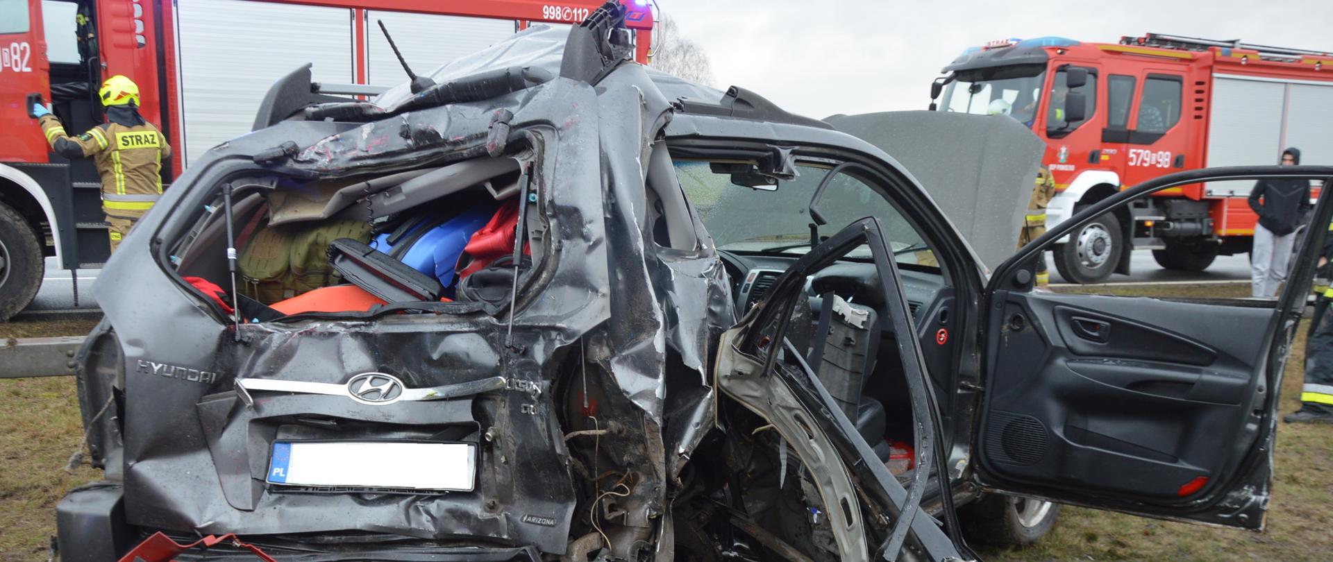 Wypadek z udziałem 5 pojazdów Na pasie zieleni oddzielającym jezdnie drogi ekspresowej S5 stoi bardzo mocno rozbity samochód osobowy koloru szarego marki hyundai tucson. Auto ma zgnieciony tył i prawą stronę oraz uszkodzony dach. W tle widoczne sa dwa wozy ratowniczo-gaśnicze oraz strażacy. 