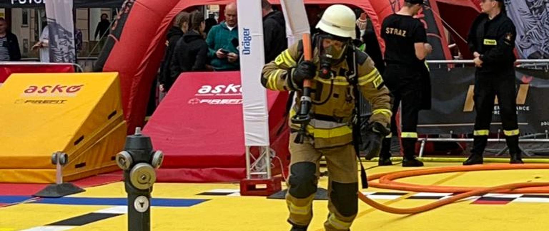 Zdjęcie przedstawia strażaka w piaskowym mundurze, białym hełmie na głowie, który biegnie po żółtej utwardzonej nawierzchni ciągnąc za sobą wąż strażacki. W tle czerwono-czarny namiot, fasady budynków oraz inni ludzie.