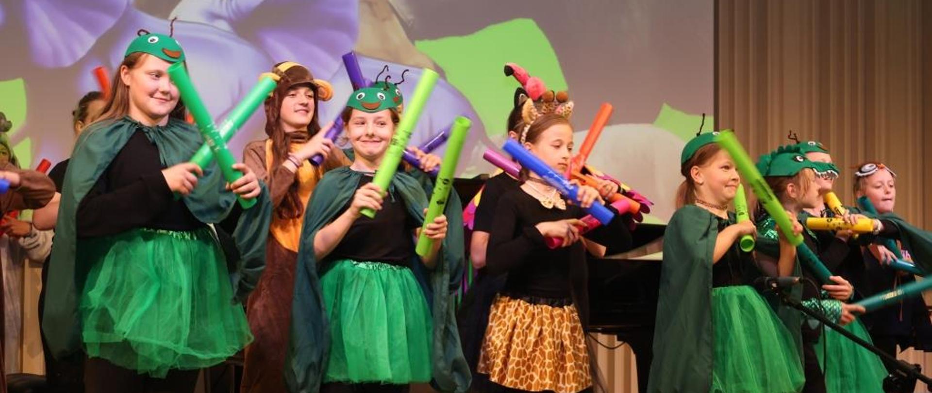 Widowisko słowno muzyczne pt. Arka Noego wita cię kolego" wg.scenariusza Beaty Makowskiej. Na zdjęciu uczniowie przebrani za zwierzęta, którzy tańczą. W tle na rzutniku grafika przedstawiająca zwierzęta. Podłoga drewniana, a tło obrazka w odcieniach beżu.