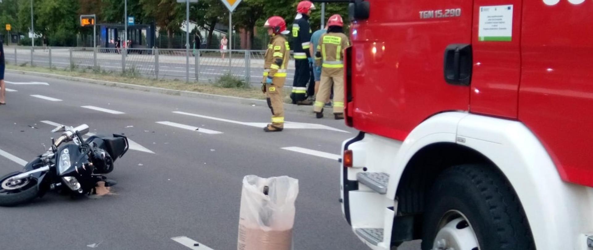 Zdjęcie przedstawia perspektywę ulicy Grunwaldzkiej, na jezdni leży uszkodzony motocykl, przed nim stoi worek Damolinu a po prawej stronie widać fragment samochodu pożarniczego i grupę strażaków przed nim.