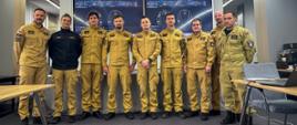 Ośmiu strażaków w beżowych mundurach pozuje do zdjęcia grupowego w sali konferencyjnej. W tle dwa monitory wyświetlające grafikę techniczną umundurowania. Jeden z uczestników nosi czarną bluzę z emblematem.