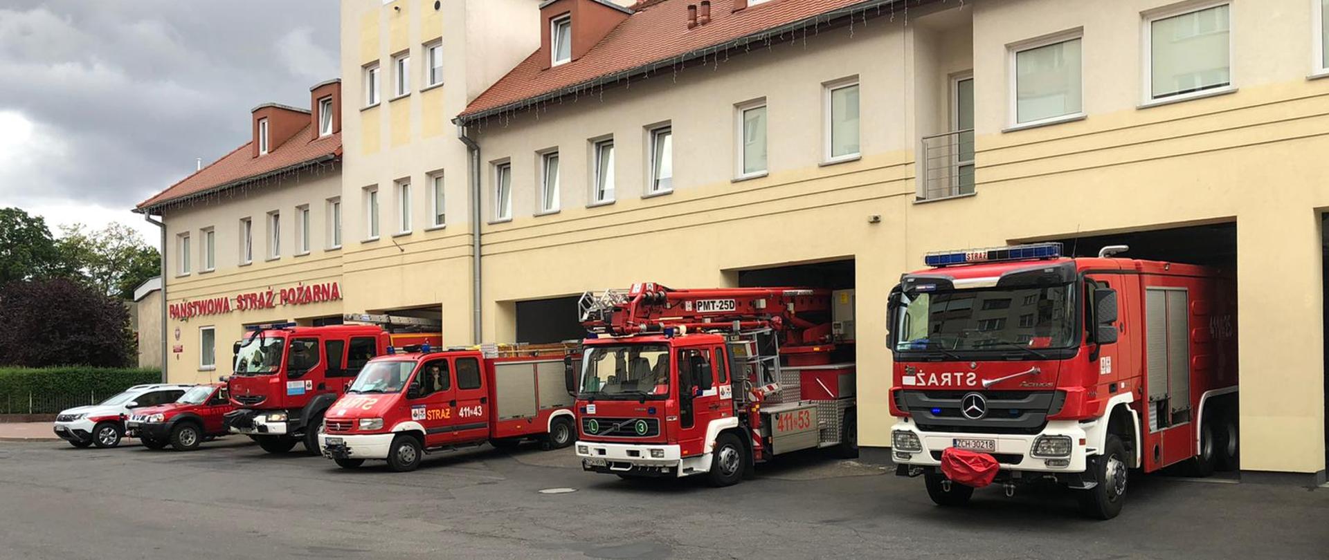 Na fotografii znajduje się sześć wozów strażackich, które wyjechały przed bramy wyjazdowe swojej jednostki. Za wozami stoi kremowy budynek Komendy Powiatowej PSP w Choszcznie. Zdjęcie wykonano w porze dziennej przy pochmurnym niebie.