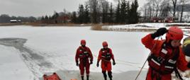 Trzech strażaków ubranych w skafandry do pracy w wodzie. Stoją na zalodzonym akwenie wodnym i przygotowują sprzęt do ćwiczeń - deska lodowa, nosze typu deska oraz linki pływające. Na drugim brzegu stawu zabudowania oraz samochód strażacki z drabiną.