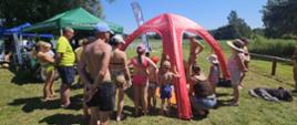 Zdjęcie przedstawia uczestników akcji „Bezpieczne Wakacje” na plaży miejskiej Ośrodka Sportu i Rekreacji w Gołdapi zgromadzonych przy stoisku edukacyjnym Komendy Powiatowej Państwowej Straży Pożarnej w Gołdapi, gdzie realizowane są również zajęcia teoretyczne i praktyczne w zakresie zasad udzielania kwalifikowanej pierwszej pomocy. Z lewej strony można zaobserwować namioty promocyjne Placówki Straży Granicznej w Gołdapi oraz Powiatowej Stacji Sanitarno-Epidemiologicznej w Gołdapi
