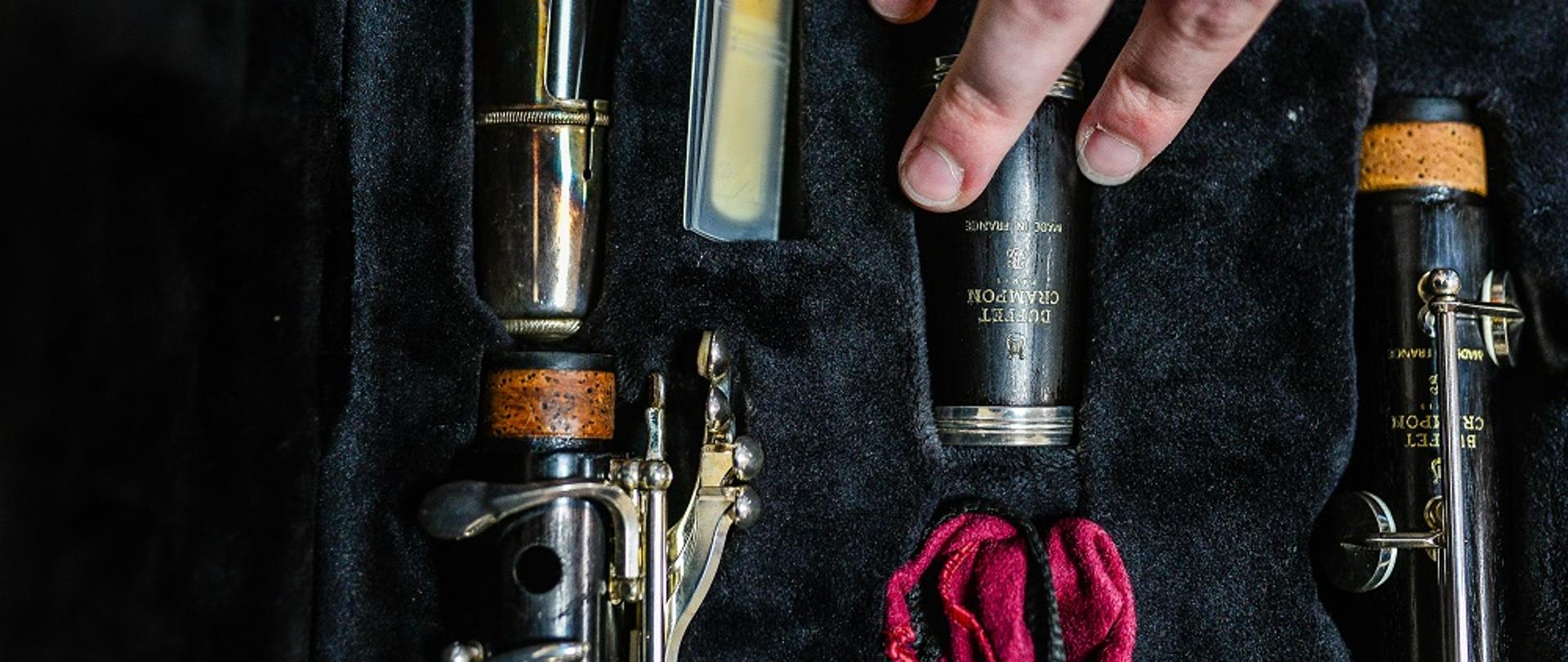 kolorowe zdjęcie przedstawia detale elementów klarnetu ułożonych w odpowiednich miejscach futerału, w prawym górnym rogu dłoń sięga po jeden z elementów instrumentu