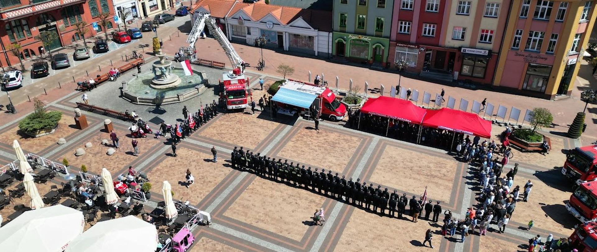 Ujęcie z drona z wysokości. Widok na płytę Starego Rynku w Chojnicach. W dwuszeregu stoją pododdziały strażaków. Na przeciwko namioty z zaproszonymi gośćmi samochód SDŁ oraz podnośnik na którym powiewa flaga Polski.