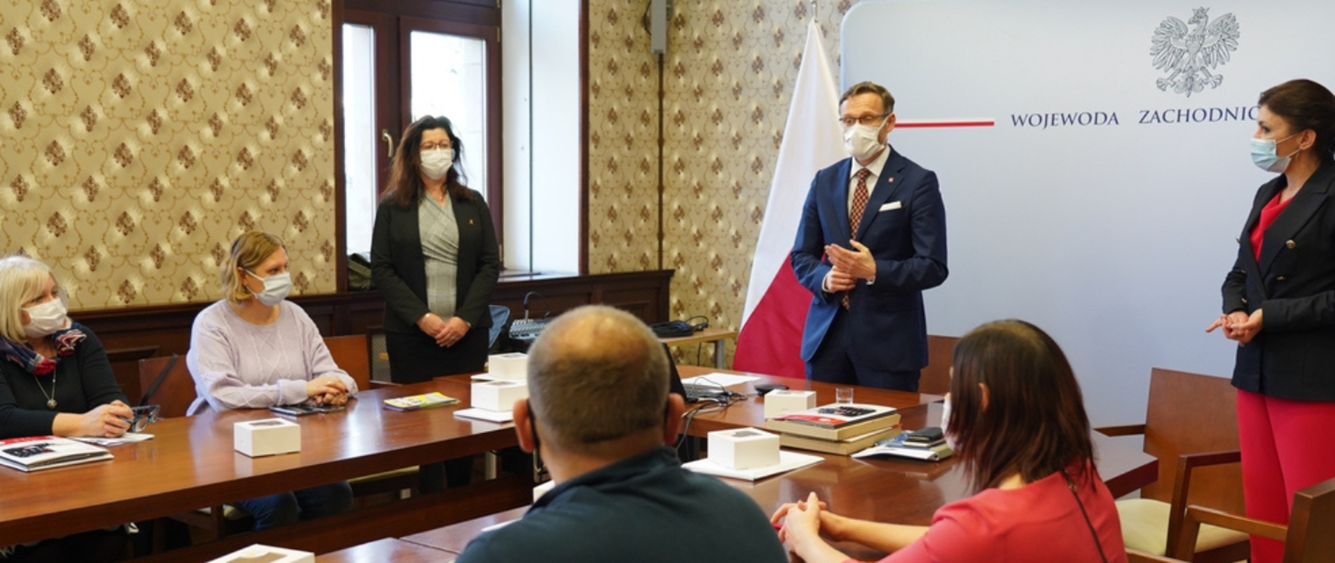 Młody mężczyzna w okularach i granatowym garniturze przemawia do słuchających go osób siedzących naprzeciw niego przy stole w kształcie litry ,,U”.