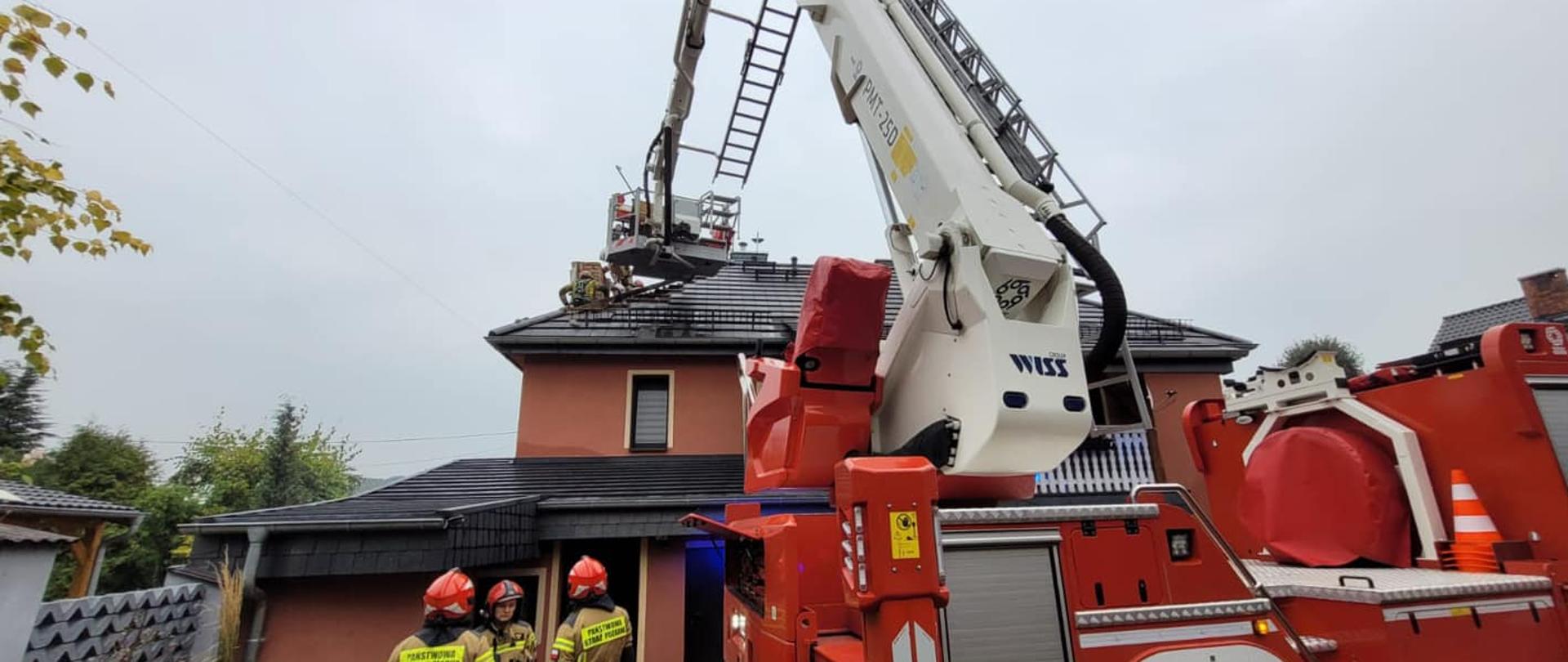 Na zdjęciu znajduje się budynek oraz strażacy, w koszu podnośnika hydraulicznego są również strażacy