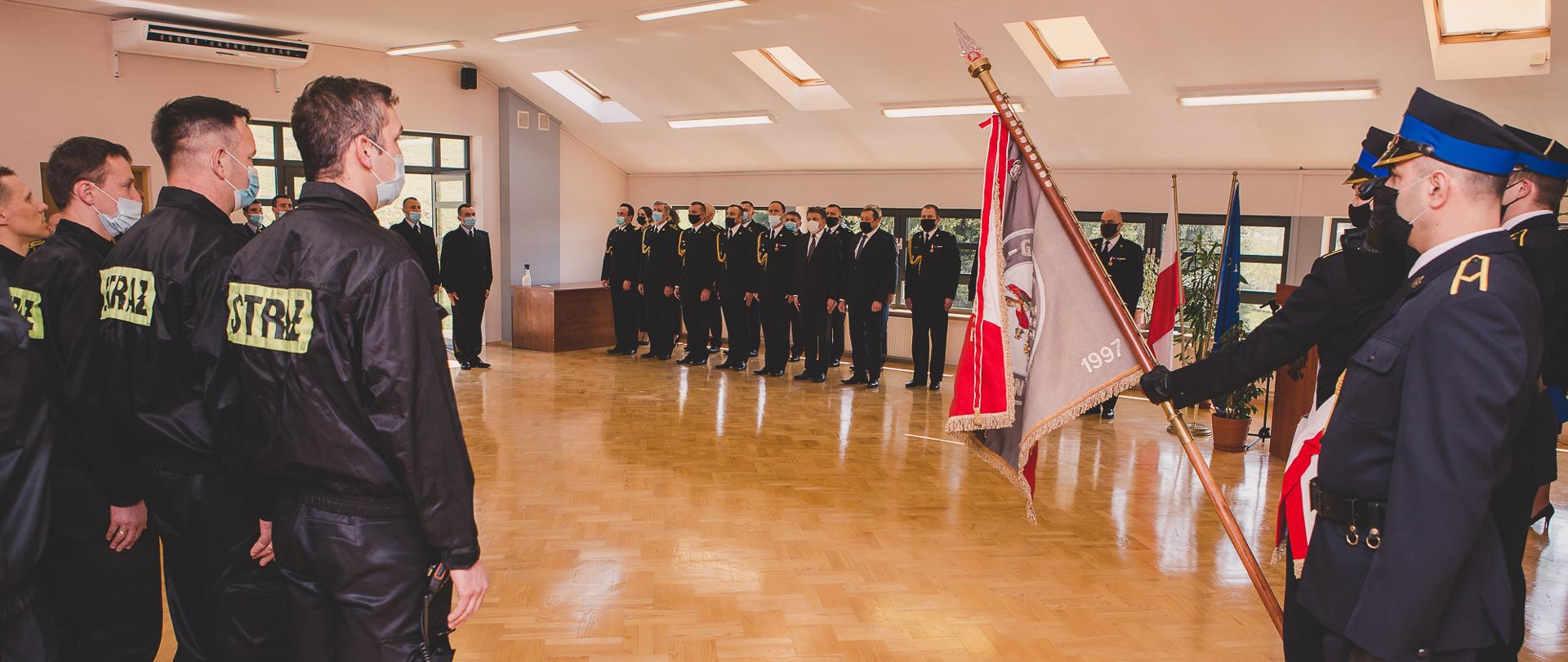 Zdjęcie wykonane wewnątrz pomieszczenia komendy. Na pierwszym planie poczet sztandarowy i kilku strażaków w umundurowaniu koszarowym, a na drugim planie pracownicy komendy i zaproszeni goście.