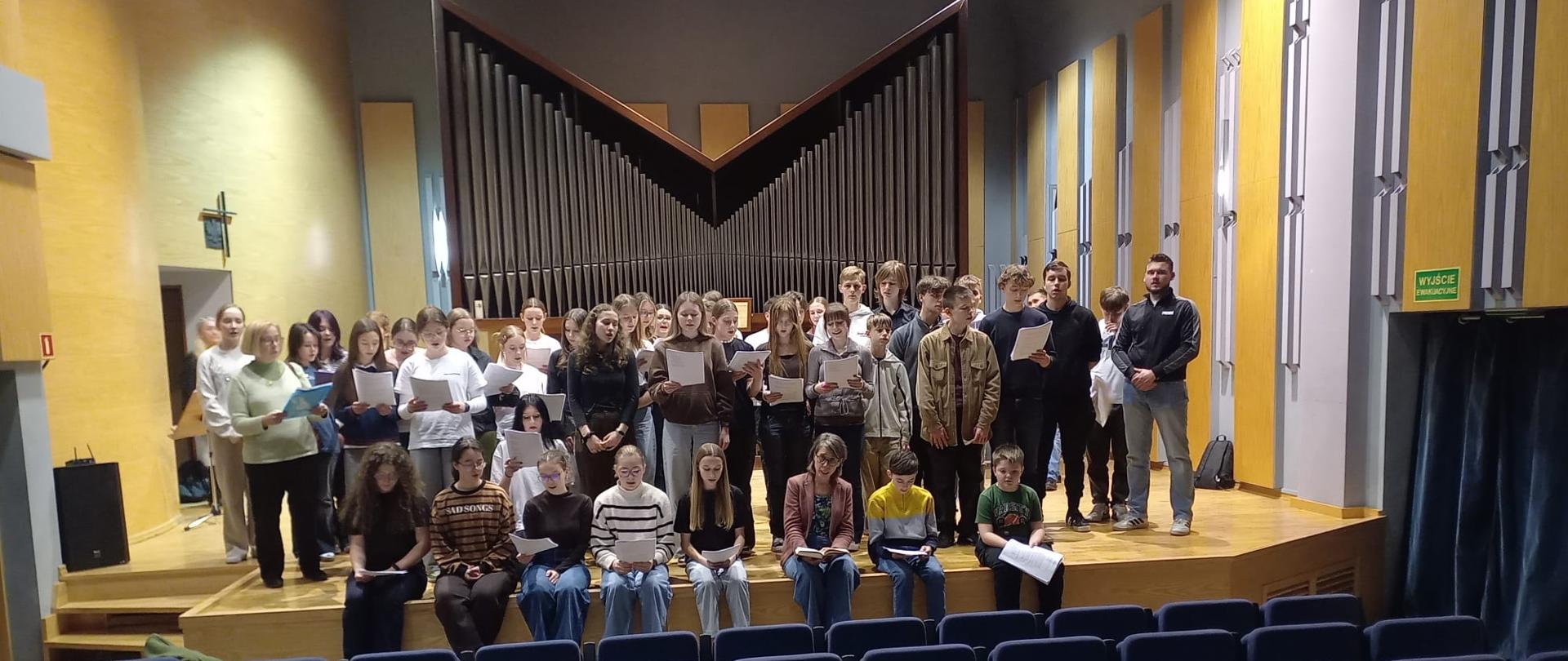 Zdjęcie przedstawia dużą grupę dzieci i młodzieży na scenie sali koncertowej, podczas warsztatów chorału gregoriańskiego. Na scenie znajduje się około 30-40 osób. Większość stoi w kilku rzędach, a kilkoro dzieci siedzi na krawędzi sceny. Akcesoria: Wiele osób trzyma w dłoniach białe kartki, co sugeruje nuty lub teksty utworów. Skupienie: Grupa wydaje się być w trakcie pracy – niektórzy patrzą w stronę nut, inni w kierunku osoby prowadzącej (niewidocznej na zdjęciu lub znajdującej się z boku) Wnętrze i scena: Organy: Najbardziej charakterystycznym elementem tła są duże, nowoczesne organy piszczałkowe o interesującym, geometrycznym kształcie litery „M” lub „V”. Sala: Wnętrze jest profesjonalne i nowoczesne. Ściany są wyłożone jasnym drewnem, co zapewnia dobrą akustykę. W dolnej części zdjęcia widać rzędy pustych, ciemnoniebieskich foteli dla publiczności. Oświetlenie: Scena jest jasno oświetlona punktowymi reflektorami sufitowymi, co kontrastuje z ciemniejszą widownią. Po lewej stronie widać wiszący na ścianie krzyż oraz profesjonalny głośnik estradowy.Zdjęcie oddaje atmosferę wspólnej pracy twórczej i pasji do muzyki. Wygląda na to, że to uczniowie szkoły muzycznej, być może właśnie tej z Nowego Targu, o której wspominałeś wcześniej.