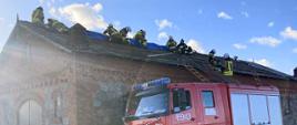 Na zdjęciu budynek z uszkodzonym dachem, na którym znajduje się ośmiu strażaków, jeden stoi na drewnianej drabinie przy krawędzi dachu, strażacy układają plandekę na uszkodzonym dachu budynku gospodarczego, przed budynkiem z czerwonej cegły stoi ciężarowy samochód ochotniczej straży pożarnej, przy budynku częściowo rozjeżdżone błoto pośniegowe, lekkie zachmurzenie z przejaśnieniami