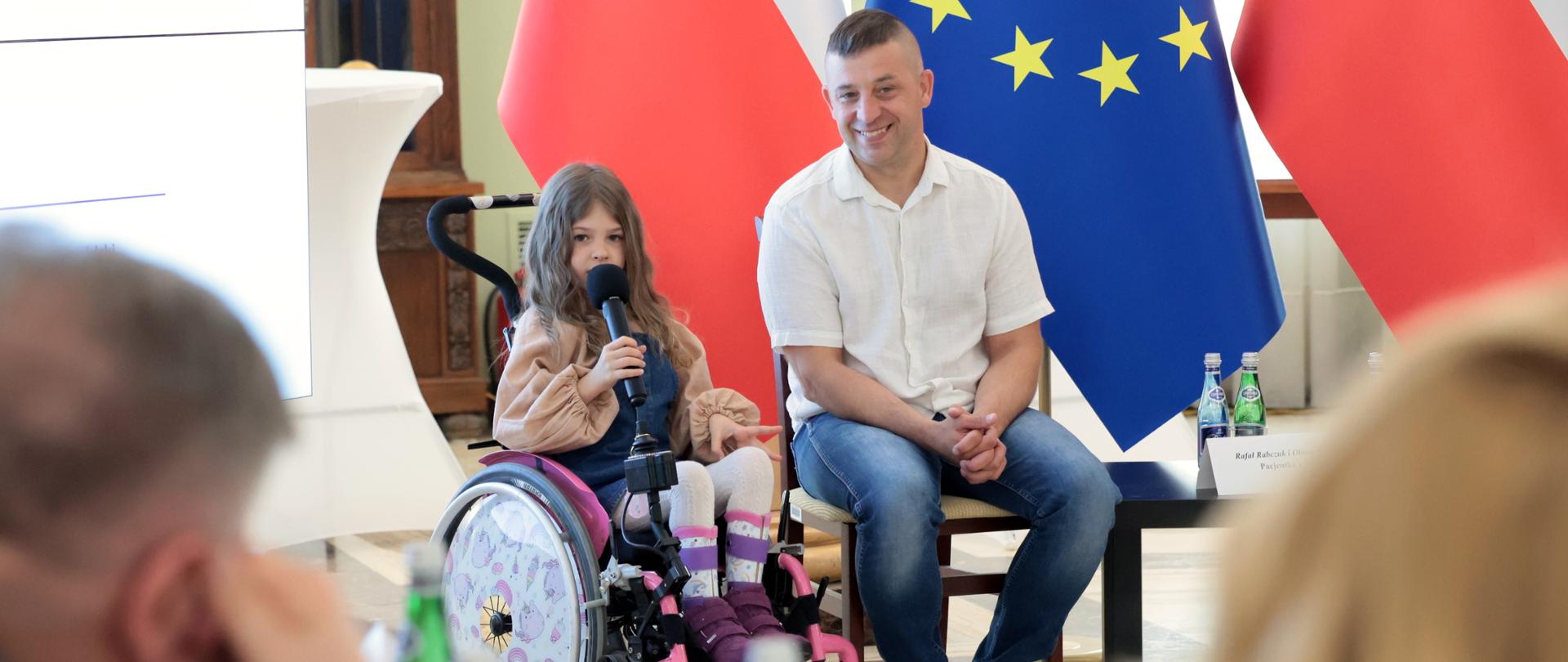 Dziewczynka siedząca na wózku inwalidzkim trzyma mikrofon i mówi. Obok niej siedzi mężczyzna i uśmiecha się. Za nimi widoczne są dwie flagi: biało-czerwona flaga Polski i niebieska flaga Unii Europejskiej z żółtymi gwiazdami. Na pierwszym planie widać fragmenty osób siedzących przy stole, a na stole stoją butelki z wodą.