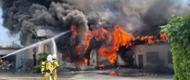 Rozległy pożar budynku gospodarczego z intensywnymi płomieniami i czarnym dymem; kilku strażaków prowadzi akcję gaśniczą przy użyciu strumieni wody.