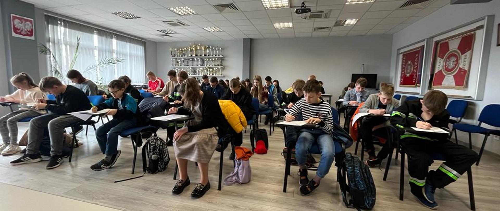 Uczestnicy turnieju piszą test. W tle gablota z pucharami.