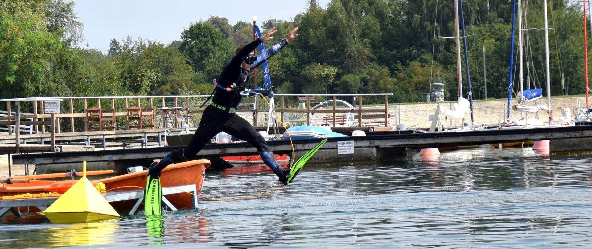 Zdjęcie zrobione na zewnątrz, przy akwenie. Na zdjęciu strażacy-nurkowie realizują zadania związane z przebiegiem mistrzostw województwa strażaków płetwonurków.
