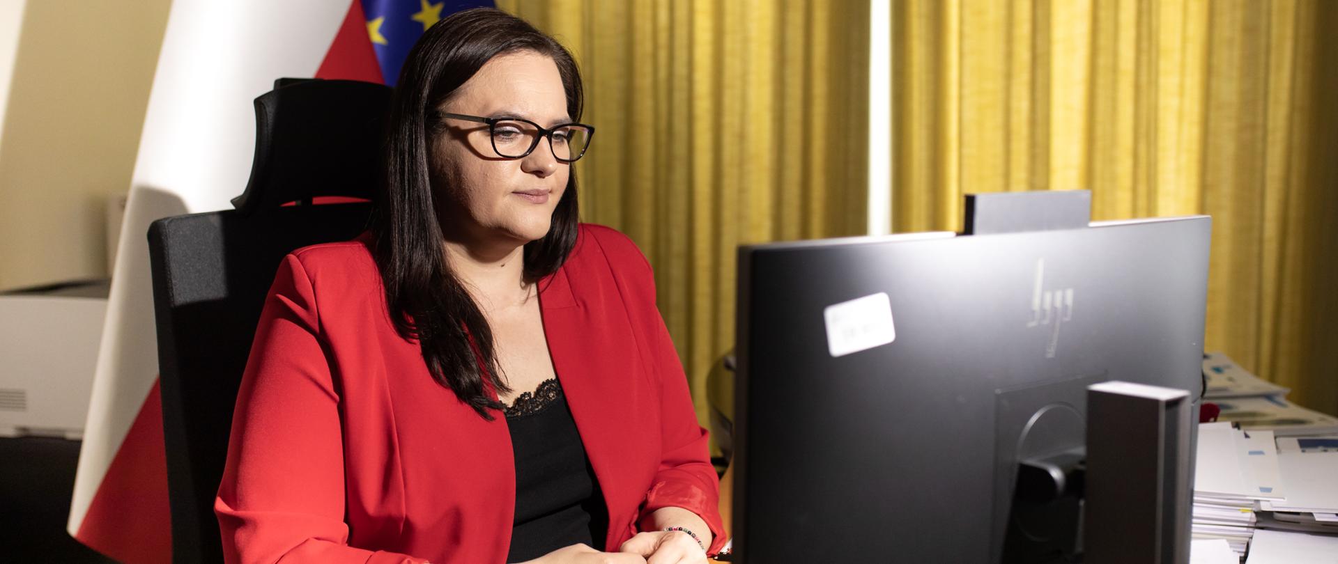 Wiceminister Małgorzata Jarosińska-Jedynak siedzi w pokoju przed monitorem. Za nią flagi PL i UE.