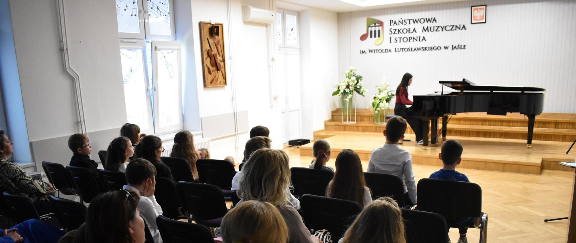 na zdjęciu, w auli PSM, na pierwszym planie widnieje społeczność szkolna, która przysłuchuje się widocznemu na drugim planie grającemu na fortepianie uczniowi. Kolorystyka zdjęcia jest biało-brązowo-szaro-czarna.