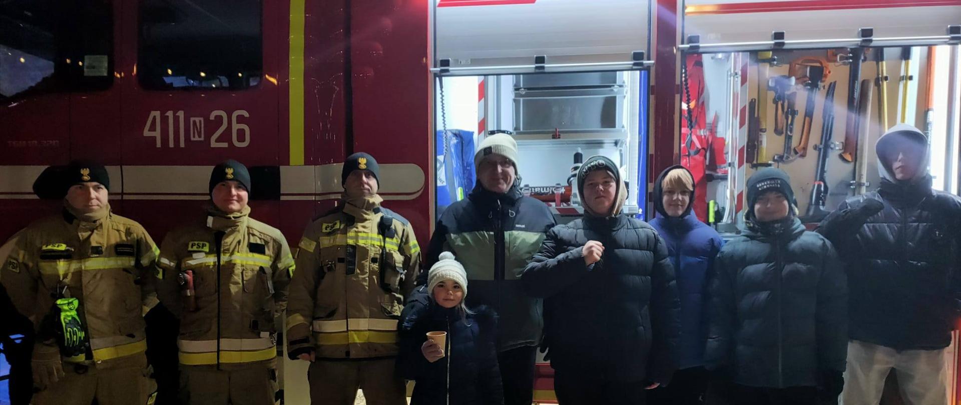 Trzech strażaków z lewej i dalej mieszkańcy Braniewa. Zima, wieczór, zimno. Za nimi samochód strażacki z otwartymi skrytkami, trawa pokaz sprzętu.