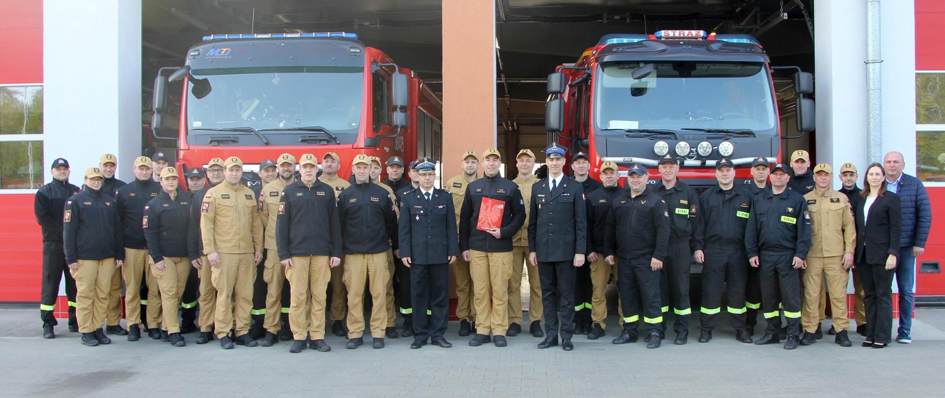 Strażacy i pracownicy cywilni stoją w dwuszeregu przed budynkiem garażowym.