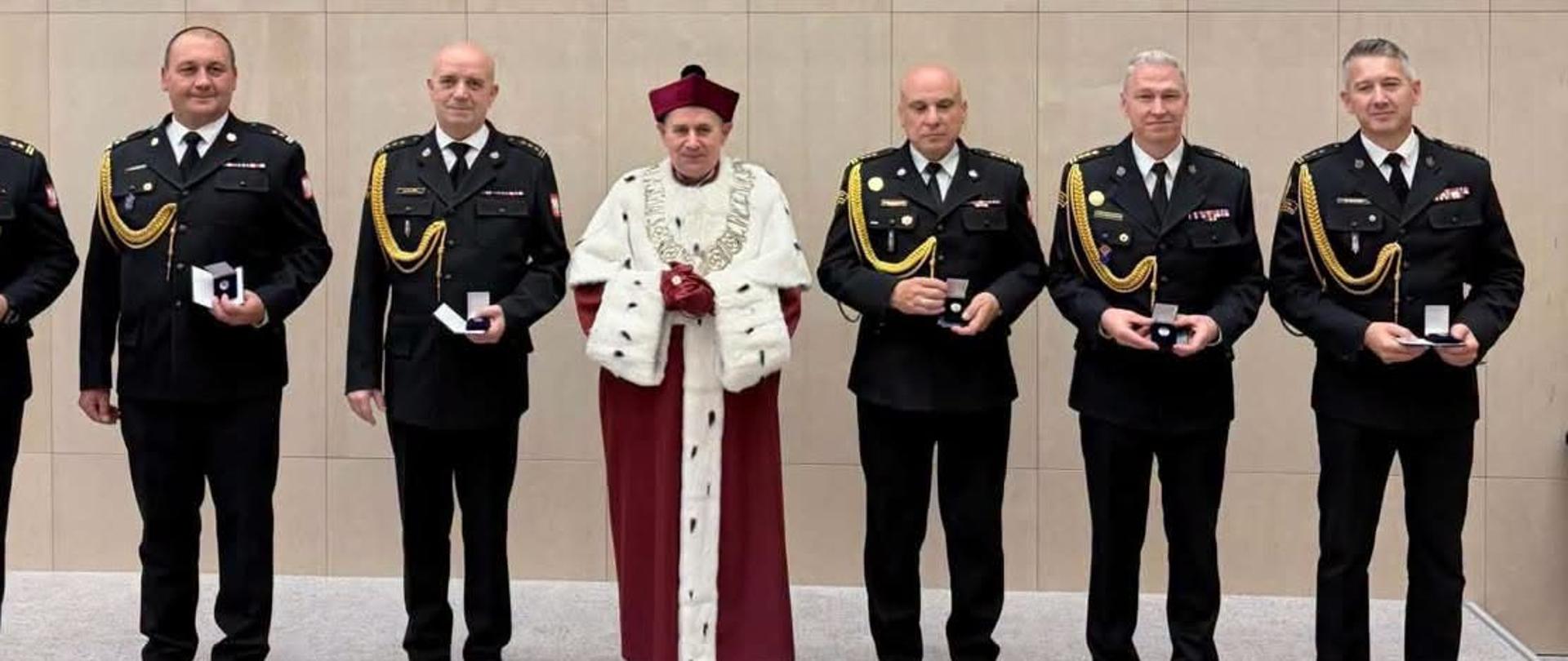 Na zdjęciu widać grupę sześciu funkcjonariuszy Państwowej Straży Pożarnej oraz duchownego, pozujących do wspólnej fotografii podczas uroczystości. Strażacy ubrani są w galowe mundury, a część z nich trzyma w dłoniach pamiątkowe odznaki. W centrum stoi rektor akademii w stroju ceremonialnym.
W tle znajduje się duża granatowa tablica z logo Śląskiego Uniwersytetu Medycznego w Katowicach oraz napisem „Odznaka Zasłużony dla Śląskiego Uniwersytetu Medycznego w Katowicach”. Na tablicy wymieniono nazwiska odznaczonych funkcjonariuszy PSP. Fotografia została wykonana w reprezentacyjnym wnętrzu uczelni.
