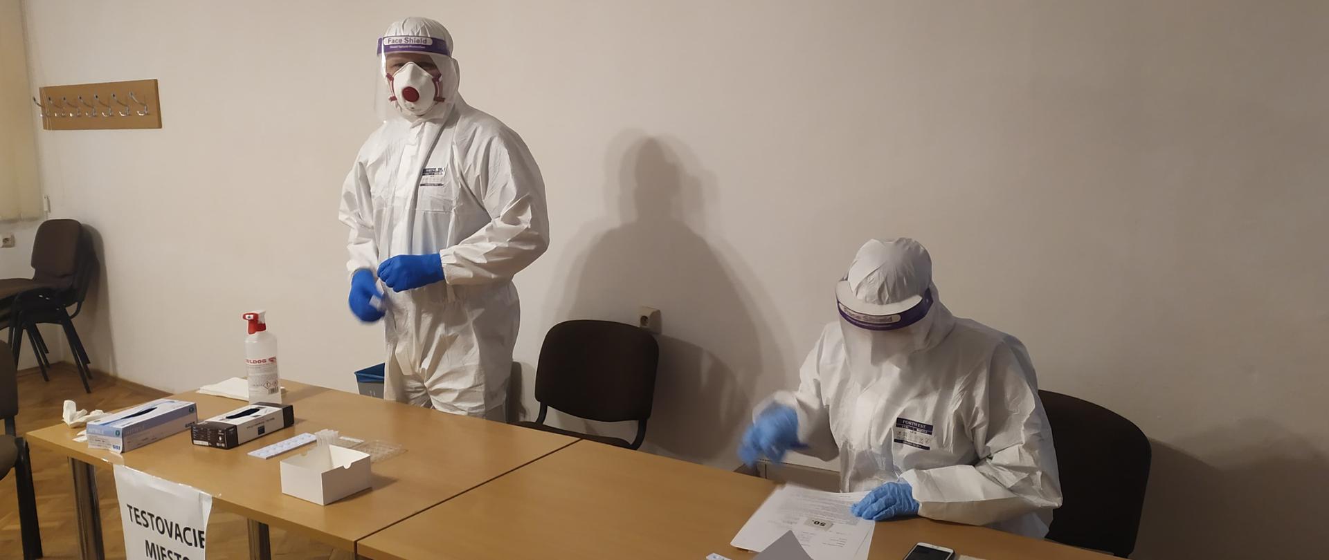 Dwie osoby zabezpieczone w kombinezony ochronne oraz maseczki w punkcie poboru wymazów.