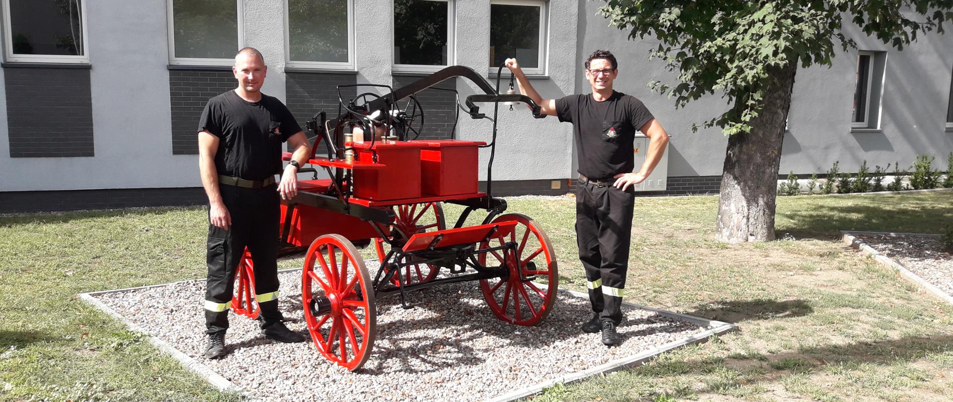 Asp. sztab. Witold Maciejewski oraz st. asp. Robert Bździak stoją przy świeżo odrestaurowanej sikawce konnej przed budynkiem wolsztyńskiej jednostki.