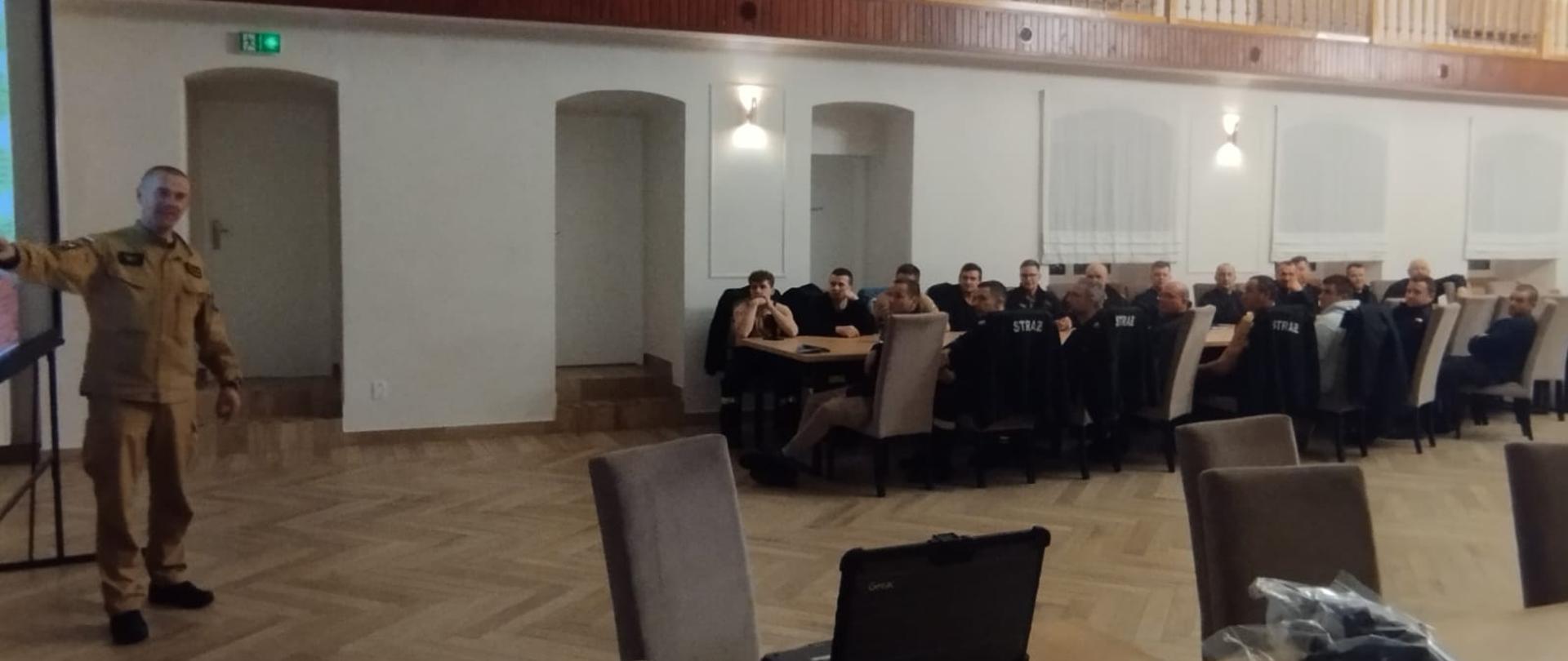 Strażak prowdzi wykład inni strażacy siedzą przy stole i słuchają.