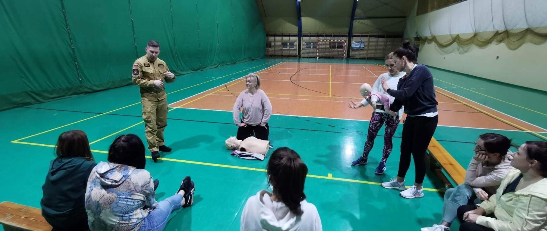 Dziewięć kobiet siedzących w okręgu, na hali sportowej, uczestniczy w szkoleniu z pierwszej pomocy "Bezpieczna Rawianka"/ które prowadzi funkcjonariusz państwowej straży pożarnej w Rawie Mazowieckiej. Strażak ubrany jest w umundurowanie służbowe w kolorze musztardowym.
