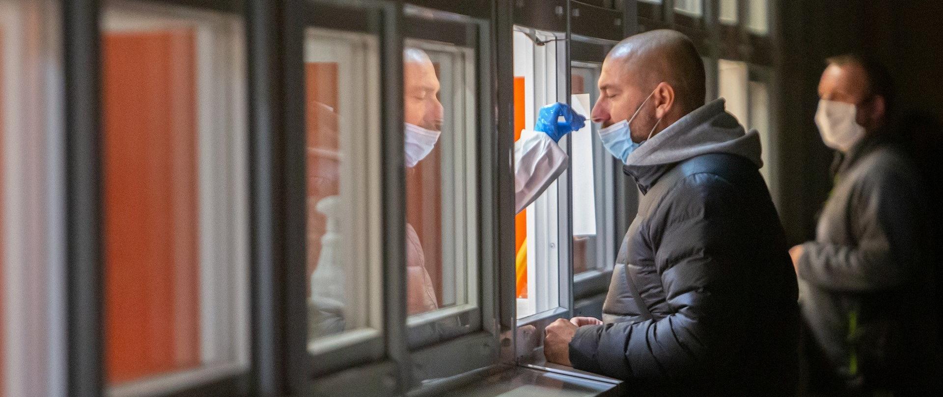 Ilustracja przykładowa Pixabay. Przy okienku stoi mężczyzna ustawiony bokiem do kamery. Jest w trakcie pobierania wymazu na obecność koronawirusa. Na zdjęciu widać również fragment dłoni, zabezpieczonej rękawiczką, która pobiera z nosa mężczyźnie wymaz. Za pacjentem w tle widać innego mężczyznę, oczekującego na badanie. 