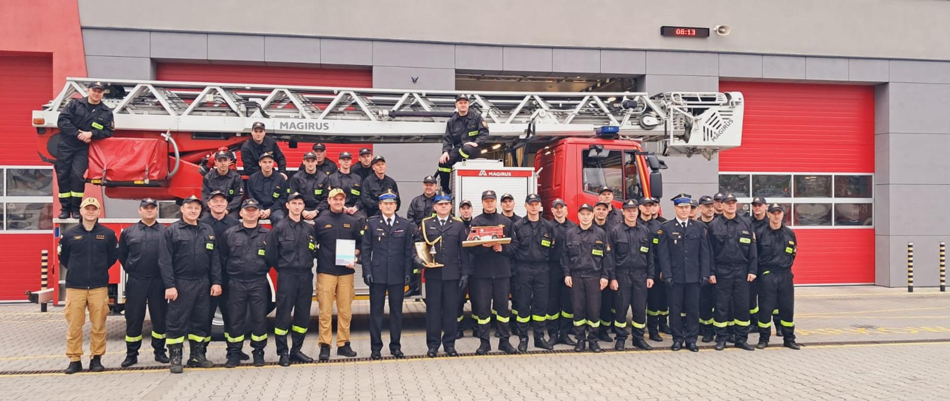 Zdjęcie pamiątkowe: wszyscy obecni na uroczystości funkcjonariusze wśród których w pierwszym szeregu stoją: komendant miejski Państwowej Straży Pożarnej w Kielcach, zastępca dowódcy Jednostki Ratowniczo-Gaśniczej Numer 1, naczelnik Wydziału Kadr i Organizacji oraz odchodzący ze służby funkcjonariusz .Tłem jest drabina mechaniczna oraz budynek JRG NR 1 w Kielcach. 