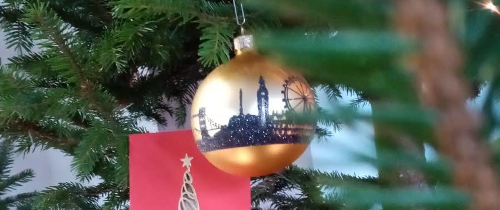 Foto przedstawia świąteczną bombkę i kartkę na tle gałęzi choinki, złota bombka z panoramą Gdańska