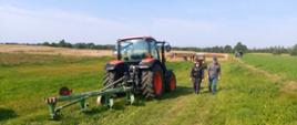 Traktor z pługiem na tle pola, w oddali grupki ludzi