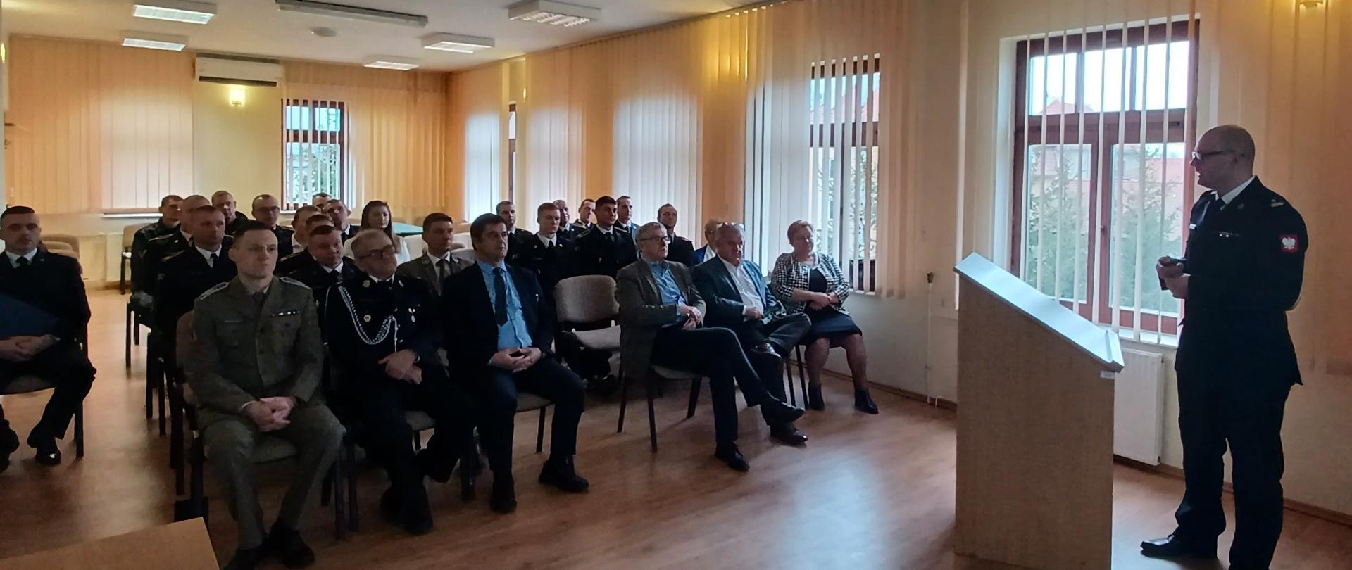 Strażacy wraz z gośćmi siedzą na świetlicy i słuchają wypowiedzi Komendanta, który stoi przed nimi. 