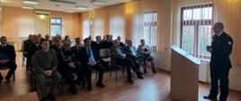 Strażacy wraz z gośćmi siedzą na świetlicy i słuchają wypowiedzi Komendanta, który stoi przed nimi. 