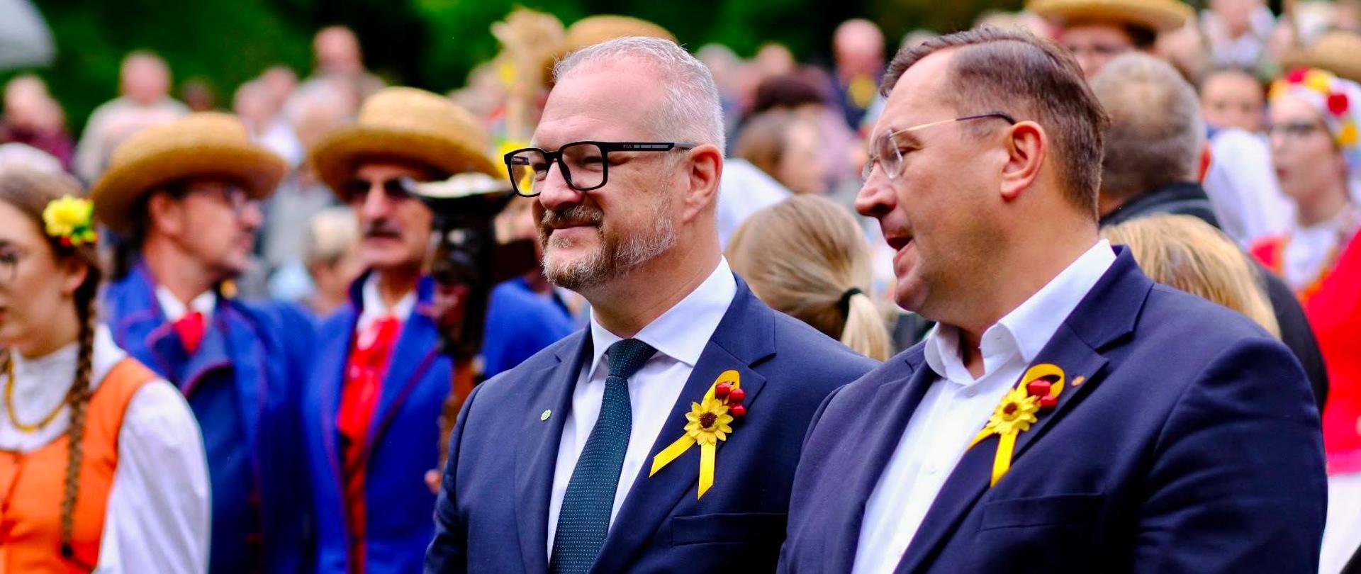 Wojewoda warmińsko-mazurski w czarnych okularach i mężczyzna w garniturze, obaj z przypinkami w kształcie słonecznika i jarzębiny, pozują do zdjęcia. Wojewoda uśmiecha się. W tle widoczni uczestnicy dożynek w strojach ludowych.
