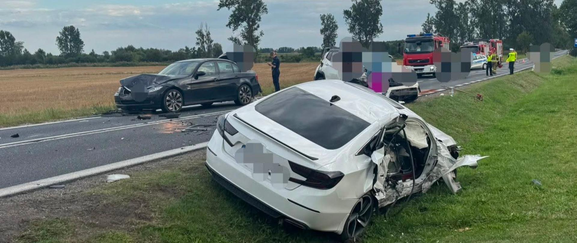 Na zdjęciu widać wypadek drogowy z udziałem kilku samochodów. Na pierwszym planie znajduje się uszkodzony biały samochód, który zjechał na pobocze, na środku drogi stoi kolejny biały pojazd z rozbitym przodem. Po drugiej stronie jezdni znajduje się ciemny samochód z uszkodzonym przodem. W tle widać wozy strażackie, radiowóz oraz służby ratunkowe zabezpieczające miejsce zdarzenia. 