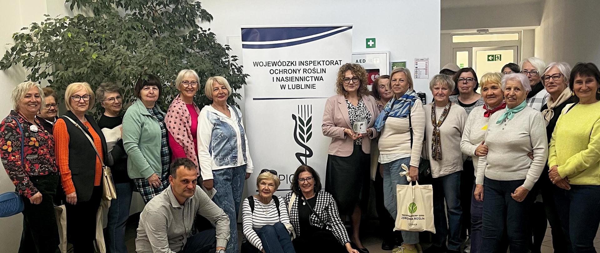 Grupa kobiet i jeden mężczyzna pozują do zdjęcia pamiątkowego. W tle duży kwiat oraz baner reklamowy.