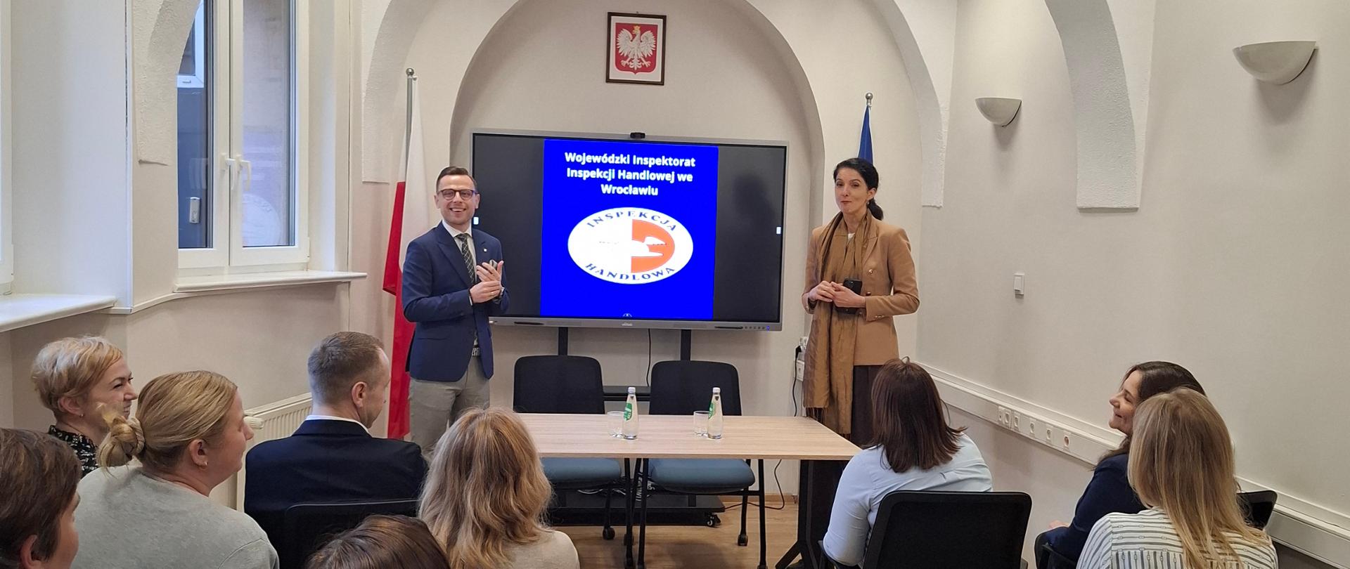 Wojewoda Dolnośląski p. Anna Żabska oraz Dolnośląski Wojewódzki Inspektor Inspekcji Handlowej p. Tomasz Resler na spotkaniu z pracownikami w Wojewódzkim Inspektoracie Inspekcji Handlowej we Wrocławiu. Pracownicy siedzą na krzesłach naprzeciwko Wojewody i Wojewódzkiego Inspektora, pomiędzy którymi wisi board z wyświetlonym logo Inspekcji Handlowej, powyżej godło Polski. 